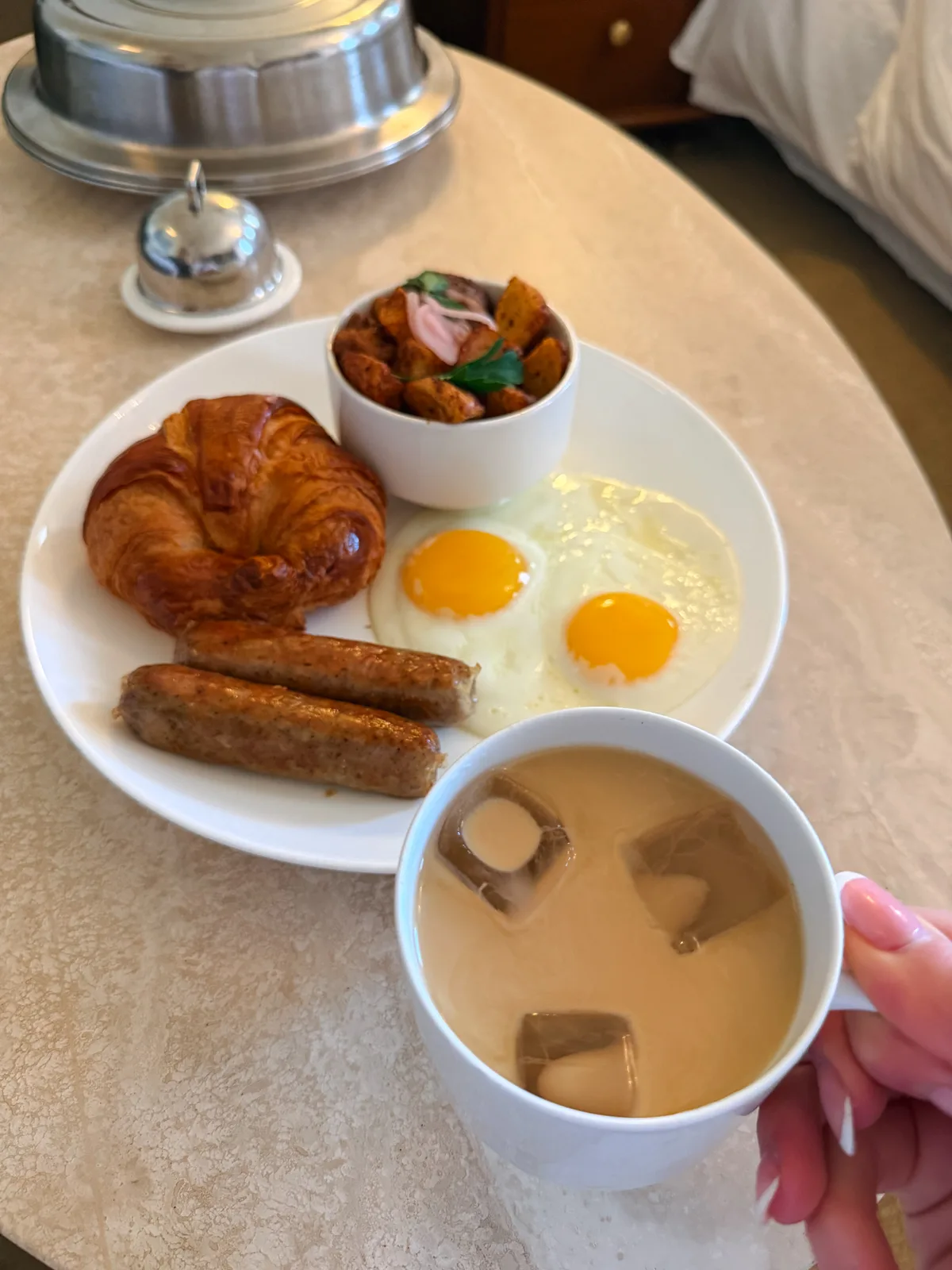 A hotel breakfast spread on a bed with pastries, coffee, and fruit in morning light
